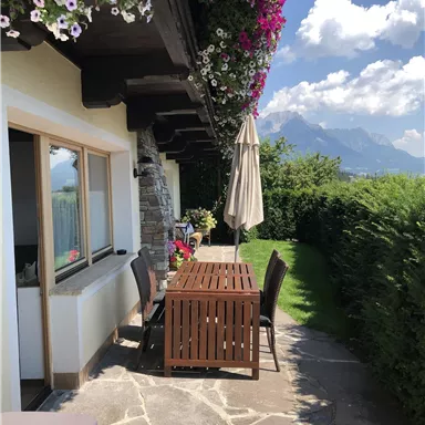 An inviting outdoor area with a table and chairs under a sunshade. Colorful flowers bloom above the table, and the mountains are visible in the background.