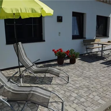 A terrace with two lounge chairs and a yellow sun umbrella. In the background, there are some blooming plants and a table with chairs visible.