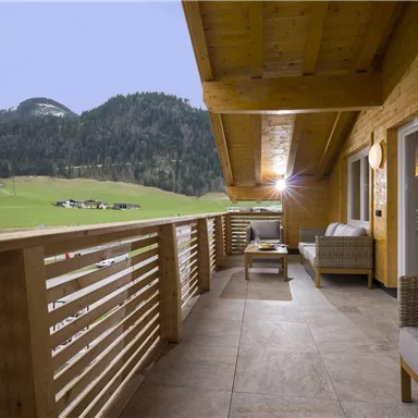 Ein Balkon mit einem Holzgeländer und gemütlichen Sitzmöbeln. Im Hintergrund sind Berge und grüne Wiesen zu sehen.