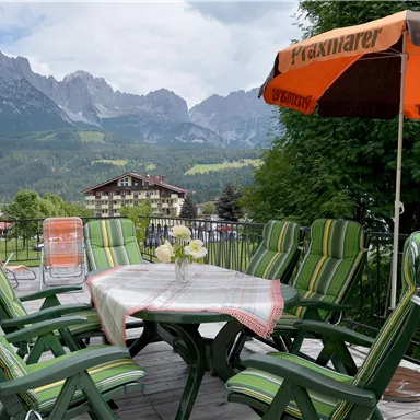 Ein einladender Terrassentisch mit grünen Stühlen und einem Sonnenschirm im Vordergrund. Im Hintergrund sind majestätische Berge und eine malerische Landschaft zu sehen.