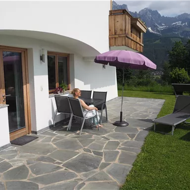 A woman is sitting on a terrace in front of a modern house. In the background, mountains and a clear sky can be seen.
