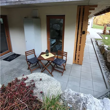 A small outdoor area with a table and two chairs. The surroundings are simple and modern, surrounded by stones and plants.