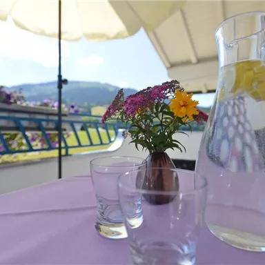 Ein schöner Tisch auf einem Balkon mit einer Glaskaraffe und zwei Trinkgläsern. Eine Vase mit bunten Blumen und eine Sonnenschirm runden die gemütliche Atmosphäre ab.