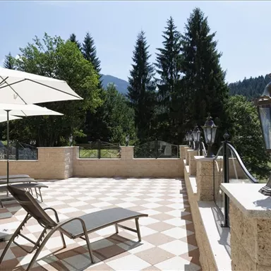 A sunny terrace with loungers and umbrellas surrounded by trees. In the background, gentle hills and a clear sky can be seen.