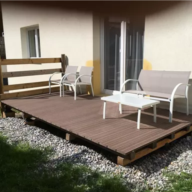 Eine kleine Terrasse mit einem Holzgeländer. Zwei Stühle und ein Tisch stehen auf dem Holzbelag, umgeben von Kies und Gras.