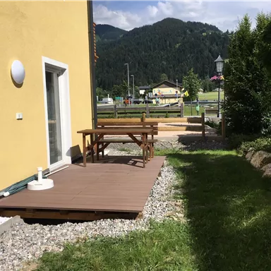 Ein sonniger Außenbereich mit einer Holzterrasse und einem Tisch. Im Hintergrund sind grüne Berge und eine gepflegte Landschaft zu sehen.