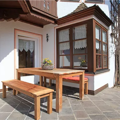 A cozy terrace with a wooden table and a bench. In the background, stacked firewood and a traditional building are visible.