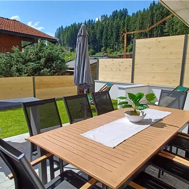 A modern terrace with a large table and chairs. In the background, there is a grill and a well-kept garden landscape.
