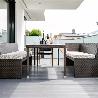 Ein gemütlicher Balkon mit einem Tisch und zwei Bänken aus Rattan. Auf dem Tisch stehen eine Flasche Wein und Gläser.