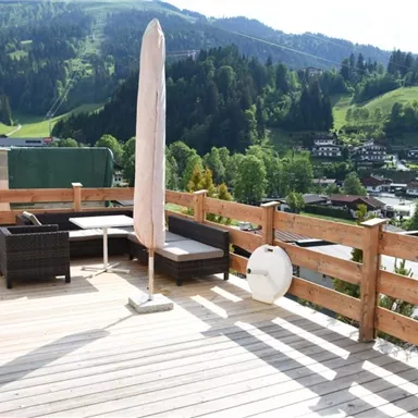 Eine gemütliche Terrasse mit Gartenmöbeln und einem Sonnenschirm. Im Hintergrund erstreckt sich eine grüne Landschaft mit Bergen und Häusern.