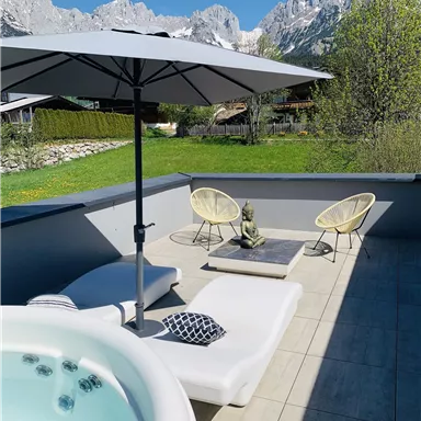 A cozy terrace with a hot tub, loungers, and a large sun umbrella. In the background, impressive mountains and a green garden are visible.