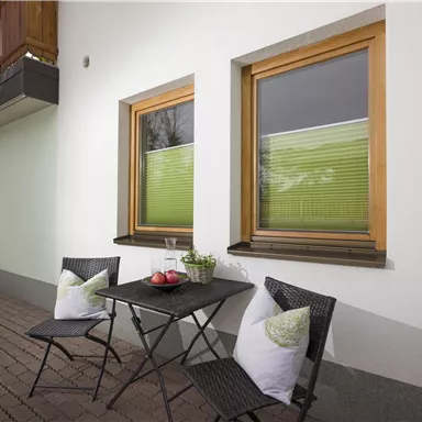 Ein kleiner, einladender Außenbereich mit einem Tisch und zwei Stühlen. Die Fenster haben grüne Rollos und die Wände sind hell gestrichen.