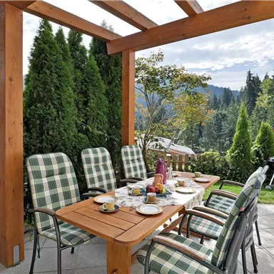 Ein schöner Essbereich im Freien, umgeben von grünen Pflanzen. Der Tisch ist festlich gedeckt und bietet einen Blick auf die Berge.