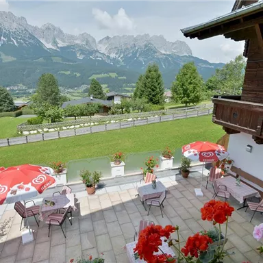 A beautiful terrace with seating and red sun umbrellas. In the background, impressive mountains and a green landscape can be seen.