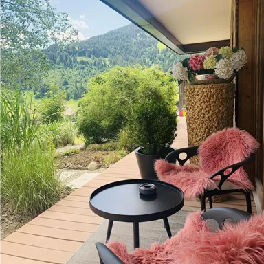 A cozy balcony with two black chairs and a small table. In the background, green mountains and decorative plants can be seen.