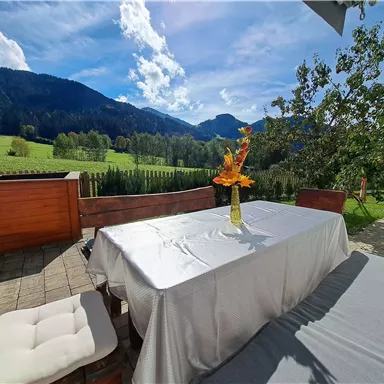 A beautiful outdoor table with a bouquet of flowers, surrounded by a green meadow and mountains in the background. The sky is blue with some clouds.