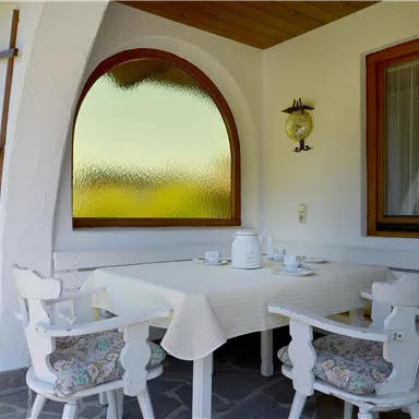 A cozy outdoor dining area with a table and chairs. The space has large windows and is surrounded by natural elements.