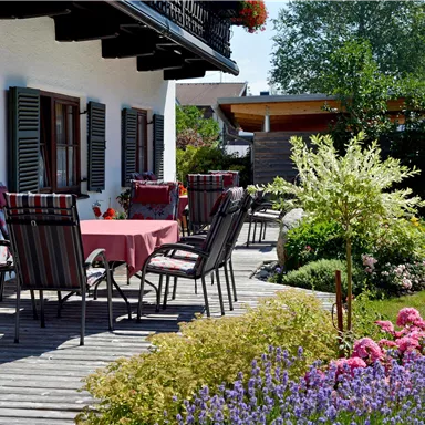 A beautiful outdoor area with a wooden table and chairs. Colorful flowers and plenty of greenery surround the terrace.