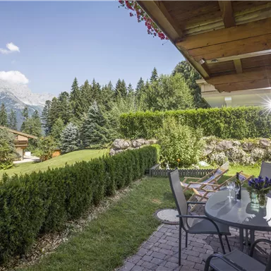 An inviting garden with well-maintained hedges and a view of the mountains. On the terrace, there are tables and chairs, ideal for relaxed hours outdoors.