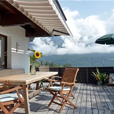A cozy terrace with wooden furniture and a sunshade. In the background, mountains and clouds are visible.