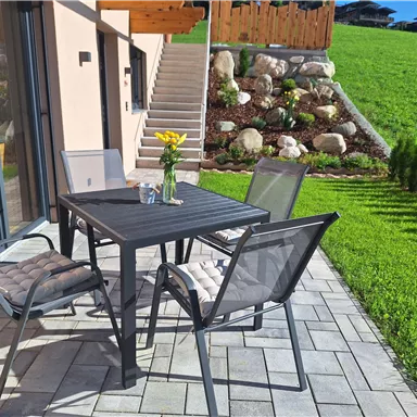 Ein gemütlicher Außenbereich mit einem Tisch und Stühlen auf einer steinernen Terrasse. Im Hintergrund grüne Wiesen und eine Treppe, die zu einem gepflegten Garten führt.