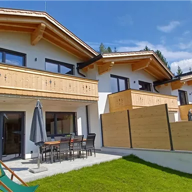 A modern house with wood cladding and a balcony. The garden is green and inviting, surrounded by mountains.