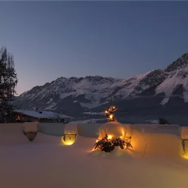 A snow-covered landscape with mountains in the background at dusk. Soft lights shine in the snow, creating a cozy atmosphere.