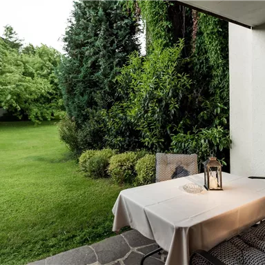 A cozy terrace with a table and chairs, surrounded by green grass and plants. The atmosphere is calm and inviting.