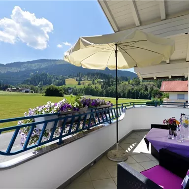 Ein schöner Balkon mit einem Sonnenschirm und blühenden Pflanzen. Im Hintergrund sind Hügel und ein blauer Himmel zu sehen.