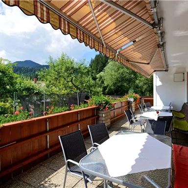 A sunny terrace with garden furniture and blooming flowers. In the background, green trees and hills are visible.