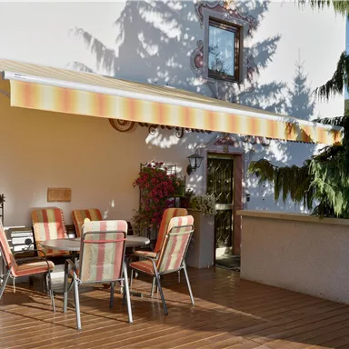 Ein gemütlicher Außenbereich mit einem Esstisch und bunten Stühlen. Über dem Tisch befindet sich ein Sonnenschirm, umgeben von Pflanzen und einem hellen Gebäude.