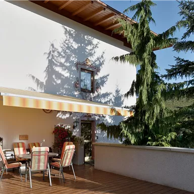 Eine gemütliche Terrasse mit einem Esstisch und Stühlen unter einem Sonnenschirm. Umgeben von grünen Bäumen und Pflanzen ist der Platz einladend und ruhig.