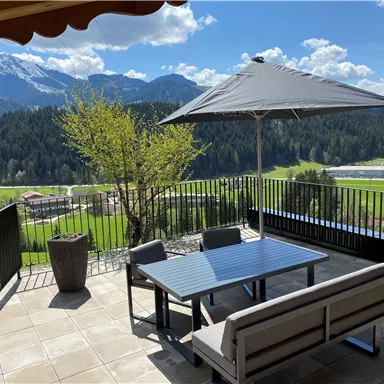 Eine gemütliche Terrasse mit Blick auf die Berge und das Tal. Der Tisch ist mit Stühlen ausgestattet und ein Sonnenschirm bietet Schatten.