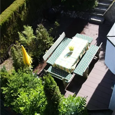 Ein gemütlicher Gartentisch mit grüner Tischdecke und Stühlen steht umgeben von Büschen. Im Hintergrund sind einige Treppen und eine gelbe Sonnenschirm zu sehen.