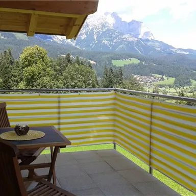 A cozy terrace with a wooden chair and table, surrounded by a yellow striped railing. In the background, a picturesque mountain landscape stretches out.