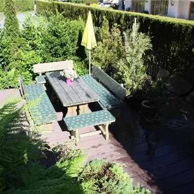 Ein gemütlicher Gartentisch mit Bänken und einem Sonnenschirm steht umgeben von grünen Pflanzen. Der Boden besteht aus Holz und wirkt einladend und frisch.