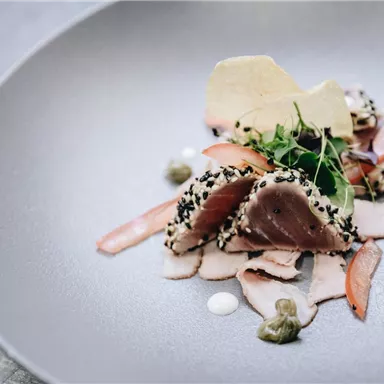 Ein kunstvolles Gericht mit Seefisch, umhüllt von schwarzem Sesam. Das Essen ist garniert mit frischen Kräutern und Saucen auf einem eleganten Teller.