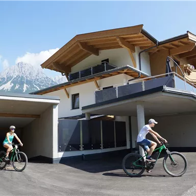 Ein modernes Haus mit Holzdekor und schönem Bergblick. Zwei Radfahrer fahren auf der Zufahrtsstraße.