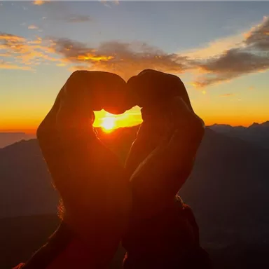 Two hands form a heart against a sunset over the mountains. The sky shines in warm colors.