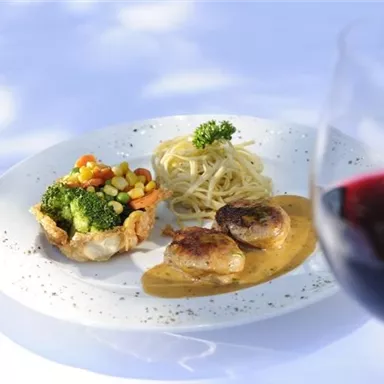 Ein köstlicher Teller mit Hähnchen, Gemüse und Spaghetti in einer Sauce. Im Vordergrund ein Glas Rotwein.