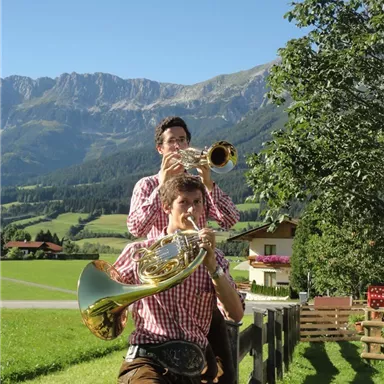 Zwei Musiker spielen in einer malerischen Berglandschaft. Die Sonne scheint und die Umgebung ist grün und einladend.