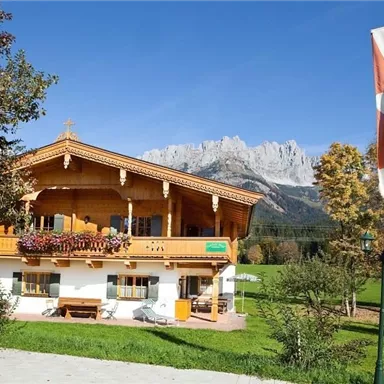 Ein traditionelles alpenländisches Holzhaus umgeben von Wiesen und Bergen. Die Fahne im Vordergrund weist auf die Region hin.