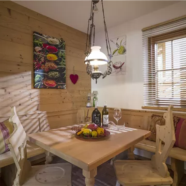 A cozy dining area with wooden furniture and warm light. On the table are a bottle of wine and a fruit bowl.