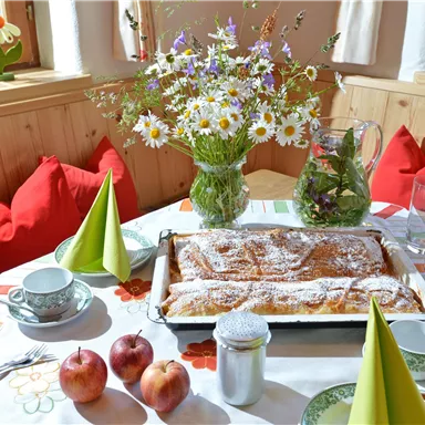 Ein einladender Tisch mit einem frisch gebackenen Apfelstrudel, Äpfeln und Teetassen. Eine Vase mit bunten Blumen und grüne Servietten runden die gemütliche Atmosphäre ab.