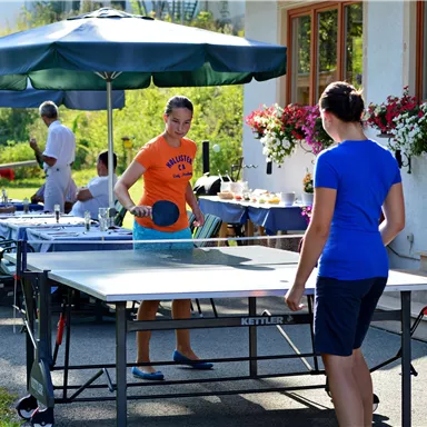 Zwei Mädchen spielen Tischtennis im Garten. Im Hintergrund sind fröhliche Gäste und blühende Pflanzen zu sehen.