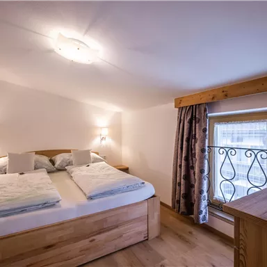 A cozy bedroom with a large bed and wooden paneling. The room has natural lighting through a window with decorative curtains.