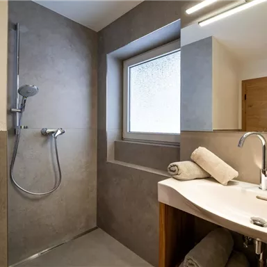 Ein modernes Badezimmer mit einer Dusche und einem Waschbecken. Die Wände sind in neutralen Grautönen gehalten, und es gibt große Fenster für natürliches Licht.