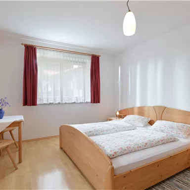 A cozy bedroom with a double bed and bright walls. There is a table by the window with a small bouquet of flowers.