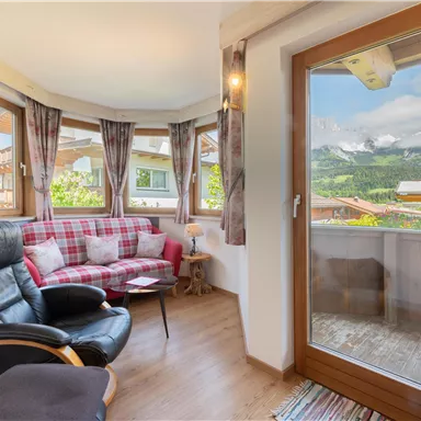 Ein gemütlicher Wohnraum mit einem grauen Sessel und einem roten karierten Sofa. Große Fenster bieten Ausblick auf die Berge und die grüne Umgebung.