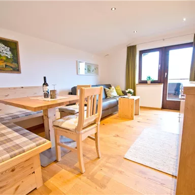 Ein gemütlicher Wohnraum mit einem Essbereich aus Holz und einer Sitzbank. Durch die großen Fenster fällt viel Licht in den Raum.